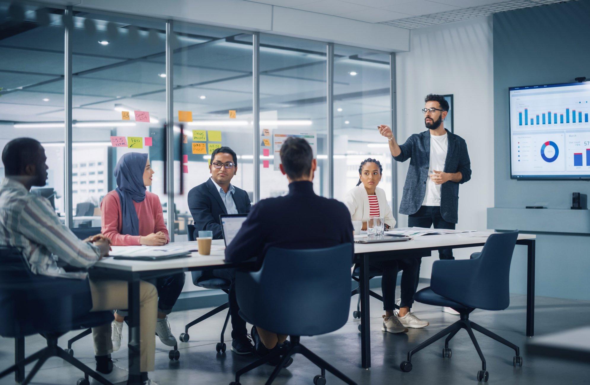 Young man presenting graphs in a boardroom to a diverse team, epitomizing 'Presenting as a Leader' skills.