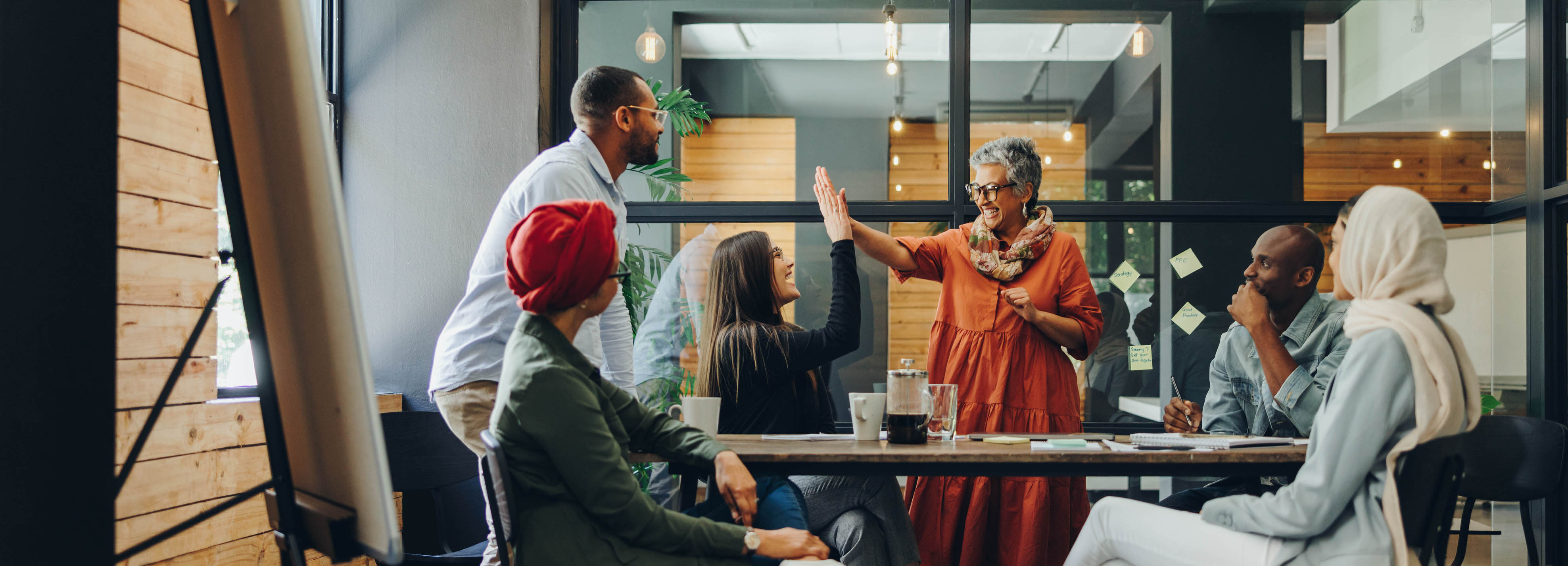 Older female leader high-fiving a team member in a diverse corporate setting, illustrating successful, authentic leadership. 