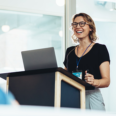 A woman in business casual attire confidently delivering a speech at a podium, representing The Humphrey Group's 'Speaking as a Leader' program.